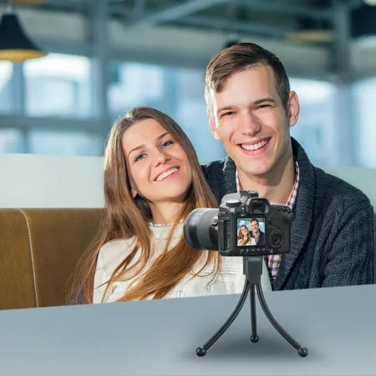 Two people sitting together with a camera on a tripod in front of them, in an indoor setting.