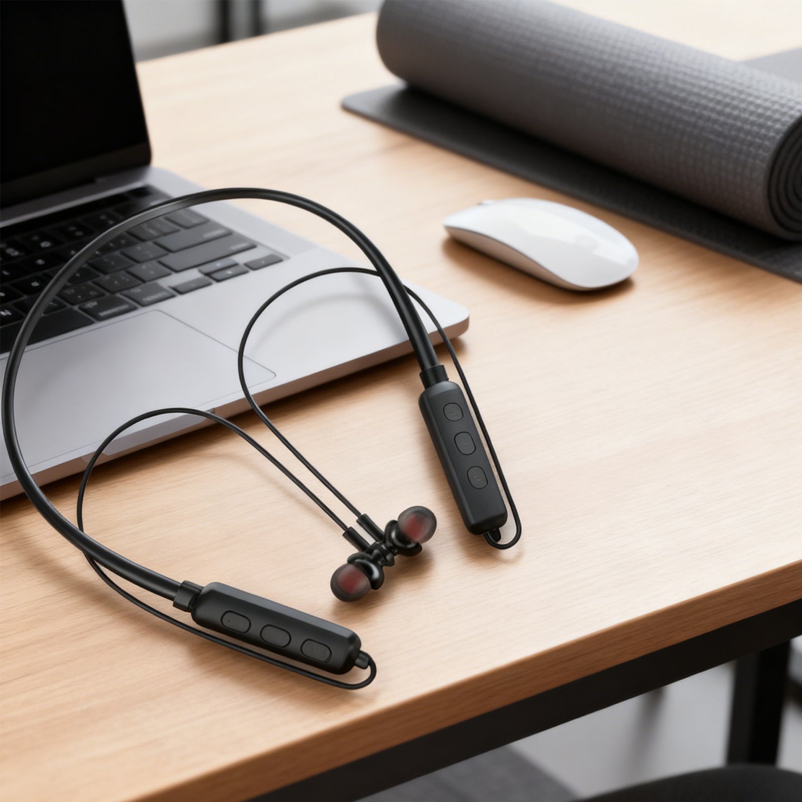 Black wireless earbuds on a desk with a laptop, mouse, and yoga mat in the background.